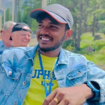 Antony Deroshan smiling in a grey baseball cap with an orange brim, a blue denim jacket, and a yellow t-shirt